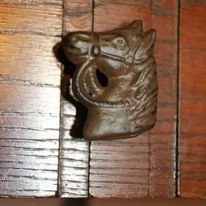 May include: A dark brown metal drawer pull shaped like a horse head with a bridle. The horse head features a textured mane and a hollow opening where the bit would be. The pull is mounted onto a horizontal wooden surface with a visible grain pattern.