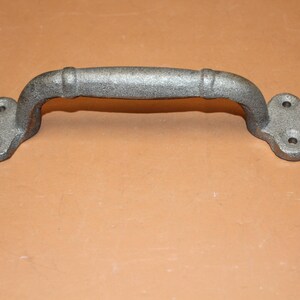May include: A gray metal door handle with a textured, cast-iron appearance. The handle features a central arched grip with two raised bands and two clover-shaped mounting plates at each end, each containing three pre-drilled screw holes.
