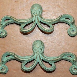 May include: Two cast iron octopus-shaped drawer pulls with a distressed turquoise finish and gold accents. The pulls have a detailed design, including the octopus's head and tentacles. These decorative pulls are ideal for nautical or coastal themed decor.