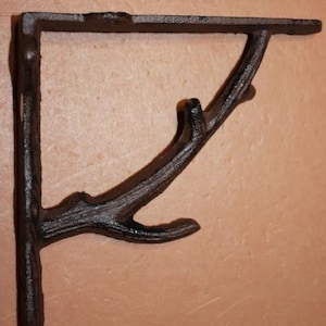 May include: A dark brown, cast iron shelf bracket with a branch-like design. The bracket has a right-angle shape with a flat top and a support that resembles a tree branch. The finish has a rustic, textured appearance.