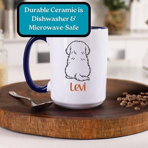 Puede incluir: Taza de cer&aacute;mica blanca con asa azul. La taza presenta un dibujo en blanco y negro de un perro sentado de espaldas al espectador. El texto "Levi" est&aacute; impreso en naranja debajo del perro. La taza est&aacute; sobre una superficie de madera con granos de caf&eacute; esparcidos a su alrededor.