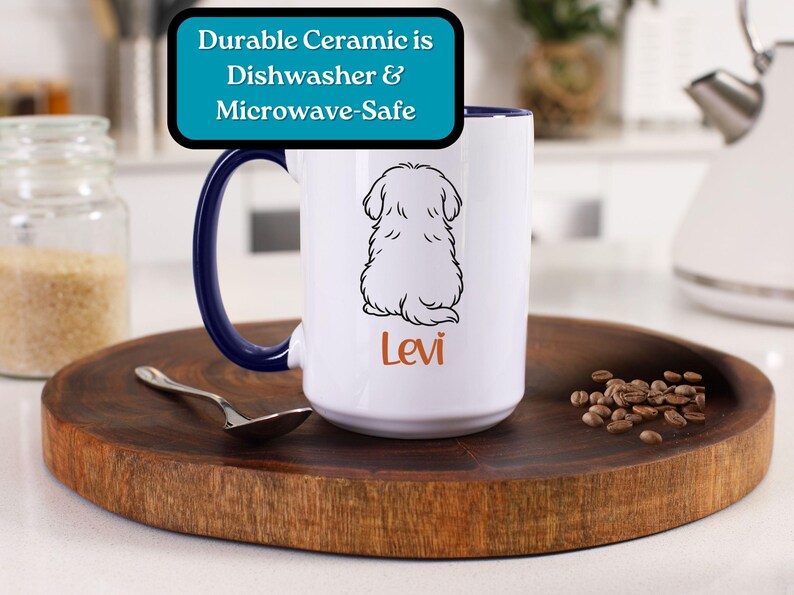 Puede incluir: Taza de cer&aacute;mica blanca con asa azul. La taza presenta un dibujo en blanco y negro de un perro sentado de espaldas al espectador. El texto "Levi" est&aacute; impreso en naranja debajo del perro. La taza est&aacute; sobre una superficie de madera con granos de caf&eacute; esparcidos a su alrededor.