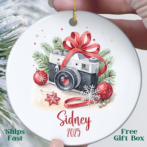 May include: A white ceramic Christmas ornament featuring a watercolor illustration of a vintage camera wrapped with a red bow and surrounded by festive decorations. The ornament includes the name "Sidney" and the year "2025".