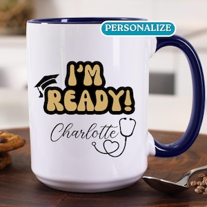 May include: White ceramic coffee mug with a blue rim. The mug has a graduation cap, stethoscope, and the text "I'm Ready!" in gold and black. The name "Charlotte" is written in black cursive below the text.