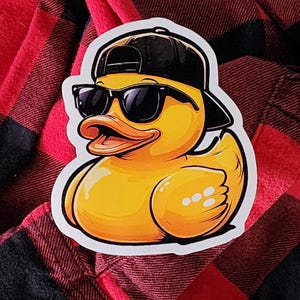 May include: A yellow cartoon duck wearing a black baseball cap and sunglasses. The duck sticker is on a red and black plaid background. The duck has an open beak and white highlights.