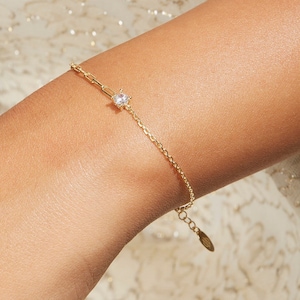 Puede incluir: Una delicada pulsera dorada con una pequeña gema transparente en el centro. La pulsera presenta una fina cadena y un pequeño eslabón rectangular cerca de la gema. La pulsera está en una muñeca, y el fondo es una tela clara y texturizada.