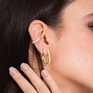 May include: Gold-toned hoop earrings and a matching ear cuff, both adorned with small, sparkling crystals. The earrings feature a unique double-hoop design, adding a touch of elegance. The jewelry is displayed on a person's ear, showcasing the pieces' delicate details.