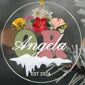 May include: A clear round Christmas ornament with a green and pink "OR" for "Our" with the name "Angela" written in cursive underneath. The letters are surrounded by dried flowers and greenery. The ornament has a white snow-like design at the bottom with the text "EST 2024" underneath.