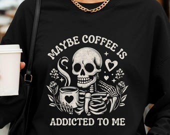 May include: Black sweatshirt with a white graphic of a skeleton holding a coffee cup with a heart. Text reads "MAYBE COFFEE IS ADDICTED TO ME". The design includes hearts and floral elements.
