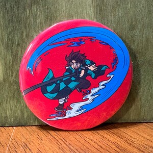 May include: A round, red button featuring a cartoon illustration of a character with a sword, surrounded by blue water-like swirls. The character is wearing a green and black checkered pattern. The button is a collectible item.