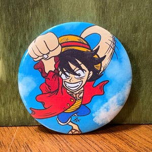 May include: A round button featuring a cartoon illustration of a character from the anime series One Piece. The character is depicted with black hair, a red jacket, and a straw hat, set against a blue sky background.