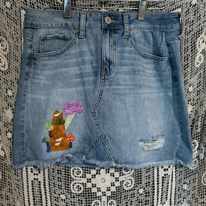 May include: A blue denim mini skirt with a frayed hem. The skirt has a patch of fabric sewn on the front, depicting a tree with mushrooms.