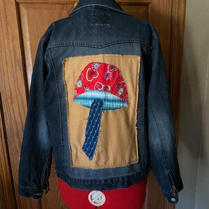 May include: A dark wash denim jacket with a large, patched mushroom design on the back. The mushroom features a red cap with heart and floral patterns, a blue and white plaid section, and a blue stem. The jacket is displayed on a red dress form.