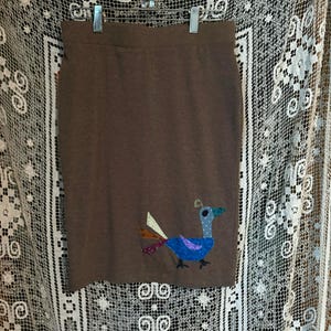 May include: A brown skirt with a colorful bird applique. The bird has a long neck, a blue body, and a yellow beak. The skirt is made of a soft, textured fabric.