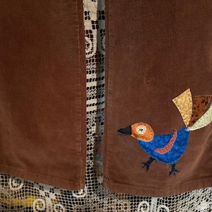 May include: A pair of brown corduroy pants with a colorful bird applique on one leg. The bird is made of fabric scraps and has a blue body, orange beak, and multi-colored tail feathers.