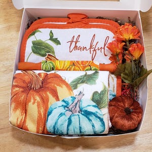 Puede incluir: Una caja de regalo llena de artículos de cocina con temática otoñal. Incluye un guante de cocina blanco y naranja y un juego de toallas a juego con diseños de calabazas y hojas. El guante de cocina tiene la palabra "thankful" en escritura naranja. También se incluyen flores decorativas naranjas y una calabaza pequeña.