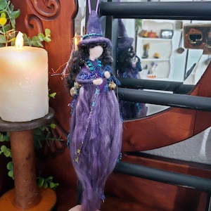 May include: A hanging witch doll ornament featuring a purple felt hat and a long, flowing purple fiber skirt. The doll has dark hair, a pale face, and is adorned with blue beaded accents and a small metallic bell hanging from its side.