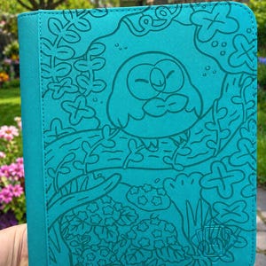 May include: Teal-colored zippered case featuring a cartoon owl design. The owl is perched on a branch surrounded by leaves and foliage. The case has a stitched border and a debossed logo in the lower right corner.