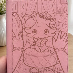 May include: A rectangular pink binder featuring a dark pink line art illustration of a small creature wearing a crown and holding a hairbrush. The creature sits on a tufted cushion surrounded by hearts, stars, and framed pictures on the wall.