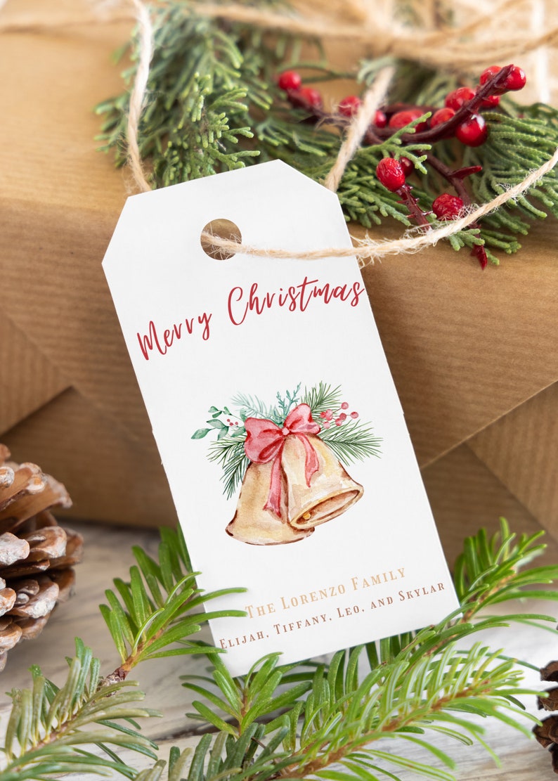 May include: White gift tag with red cursive text "Merry Christmas" and a watercolor illustration of two gold bells with a red bow and green pine branches. The text "The Lorenzo Family Elijah, Tiffany, Leo, and Skylar" is printed in gold at the bottom of the tag.