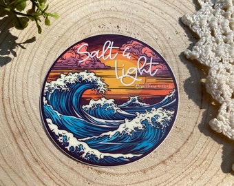 Puede incluir: Pegatina redonda con un diseño de olas estilizadas en tonos azules, blancos y morados, con un fondo de puesta de sol. El texto "Salt & Light" está escrito en blanco, con "Matthew 5:12-16" debajo.