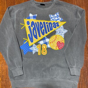 May include: A gray sweatshirt with a yellow pennant that says "Javelinas" in blue letters. The pennant is surrounded by blue and white stars and a yellow circle with a red heart and the number 18.
