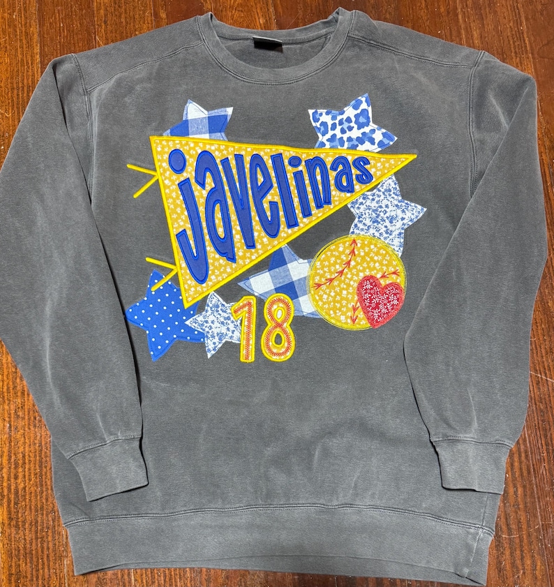 May include: A gray sweatshirt with a yellow pennant that says "Javelinas" in blue letters. The pennant is surrounded by blue and white stars and a yellow circle with a red heart and the number 18.