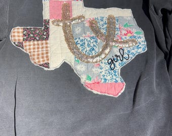 Könnte beinhalten: Ein graues Sweatshirt mit einem Patchwork-Design des Bundesstaates Texas. Das Design enthält ein Pailletten-Hufeisen und das Wort "girl" in schwarzer Stickerei.
