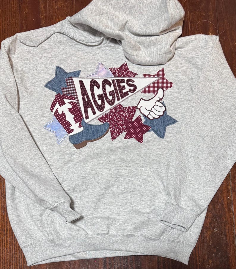 May include: A gray hooded sweatshirt with a colorful applique design featuring a pennant with the word "Aggies" and a cowboy boot. The design also includes stars and a thumbs up.
