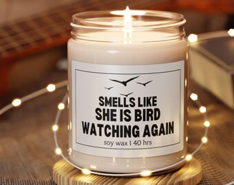 May include: A lit candle in a clear glass jar with a label that reads "Smells Like She Is Bird Watching Again." The candle sits on a wooden coaster and is surrounded by string lights. The candle is made of soy wax and has a 40-hour burn time.