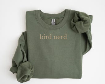 May include: An olive green sweatshirt with the words "bird nerd" printed in a light brown color across the chest. The sleeves are folded, and the sweatshirt is made of a soft material. The sweatshirt is a casual style.