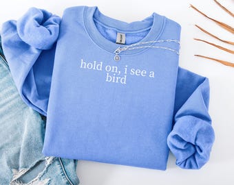 May include: A periwinkle blue sweatshirt with the phrase "hold on, i see a bird" printed in white. A silver necklace with a circular pendant rests on the shirt. The sweatshirt is folded next to a pair of blue jeans.