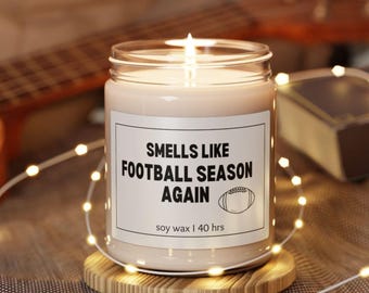 May include: A lit soy wax candle in a glass jar with a label that reads "Smells Like Football Season Again" and a football graphic. The candle sits on a wooden coaster and is surrounded by string lights. It has a 40-hour burn time.