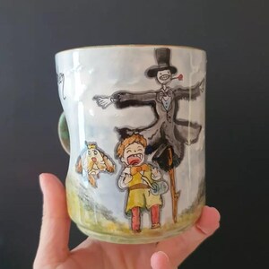 Może przedstawiać: Ceramiczny kubek z ręcznie malowaną ilustracją postaci z filmu fantasy. Projekt zawiera stracha na wróble w garniturze, młodego chłopca w żółtym i stworzenie przypominające psa. Kubek ma zielony uchwyt.