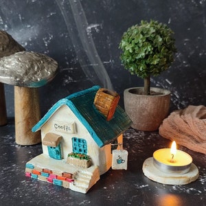 May include: A white ceramic house with a blue roof and a sign that says "Coffee". The house has a small window, a door, and a chimney. There is a small potted plant in front of the house. The house is sitting on a black surface with a small candle in front of it.