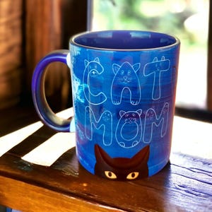 Puede incluir: Taza de cerámica azul con un contorno blanco de gatos que forman las palabras "Cat Mom". La taza tiene un gato negro asomándose desde la parte inferior.