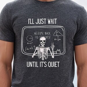 May include: A dark heather grey t-shirt with a skeleton wearing a cap and gown holding a chalkboard that says "Welcome back to students!" and has math equations on it. The text on the shirt says "I'll just wait until it's quiet."