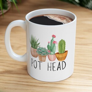 May include: White ceramic coffee mug with a black coffee inside. The mug has a graphic of five potted succulents with the text "POT HEAD" printed below.