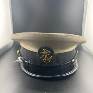 May include: A khaki-colored service cap featuring a black band, a black visor, and a gold-toned metal anchor emblem centered on the front. The cap includes a thin black chin strap secured by small gold buttons on each side of the structure.