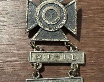 May include: A silver military marksmanship badge with a target in the center and the words "RIFLE" and "PISTOL" on two bars hanging below.
