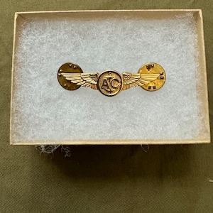 May include: A gold-colored military pin with wings, the letters "AC", and an anchor emblem. The pin is displayed inside a small, open cardboard box filled with white padding, set against a green background.