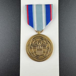 May include: A circular bronze-colored medal featuring an eagle with outstretched wings above a shield, set against a globe design with curved lines. The medal is suspended from a ribbon with vertical stripes of blue, white, and red.