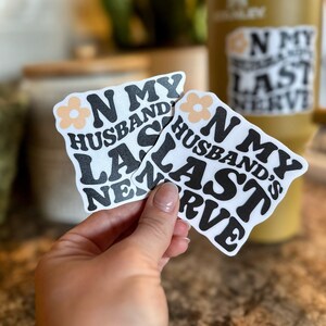 May include: Two white stickers with black text that reads "ON MY HUSBAND'S LAST NERVE" with a peach flower accent. A yellow insulated tumbler with a matching sticker is in the background.