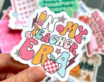 May include: A colorful sticker with the text "WOMY TEACHER ERA" in a retro font. The sticker features a pencil, a star, a smiley face, and a red and white checkered apple.
