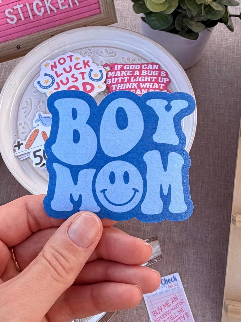 May include: A blue sticker with the words "BOY MOM" in a retro font, featuring a smiling face in place of the "O" in MOM. The sticker is held by a hand, with other stickers and a pink letter board in the background.
