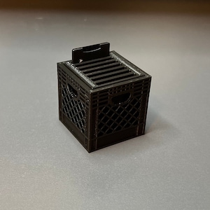 May include: A black plastic crate with a slot on top for holding items. The crate is approximately 10 cm tall and 10 cm wide.