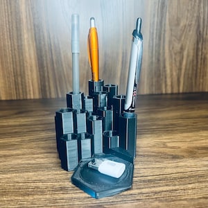 Könnte beinhalten: Schwarzer Plastik-Stiftständer mit Wabenmuster. Der Ständer hat drei Stifte darin: einen orangen, einen schwarzen und einen silbernen. Der Ständer steht auf einer Holzoberfläche.