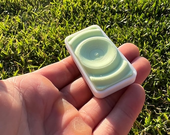 May include: A small, white and light green rectangular object with a raised, circular design. The object has a smooth, glossy finish.