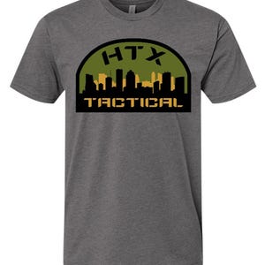 Könnte beinhalten: Graues T-Shirt mit dem Text "HTX TACTICAL". Die Grafik zeigt eine Silhouette einer Stadtlandschaft in Schwarz und Gold vor einem grünen und schwarzen Bogenhintergrund.