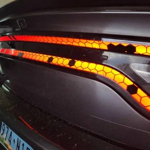 May include: Close-up of a car's taillight assembly. The taillight features a honeycomb pattern with orange and red illumination. The car's body is dark gray.